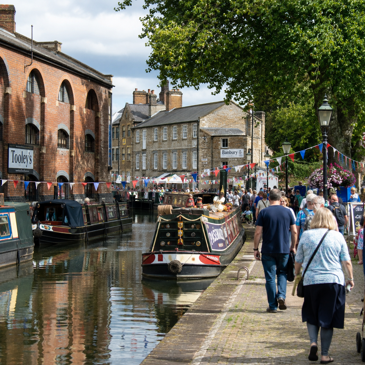 Banbury Canal Festival a busy canal-side street scene with narrowboats docked along the water, pedestrians walking on the paved embankment, brick buildings on both sides, hanging bunting, and green trees overhead.