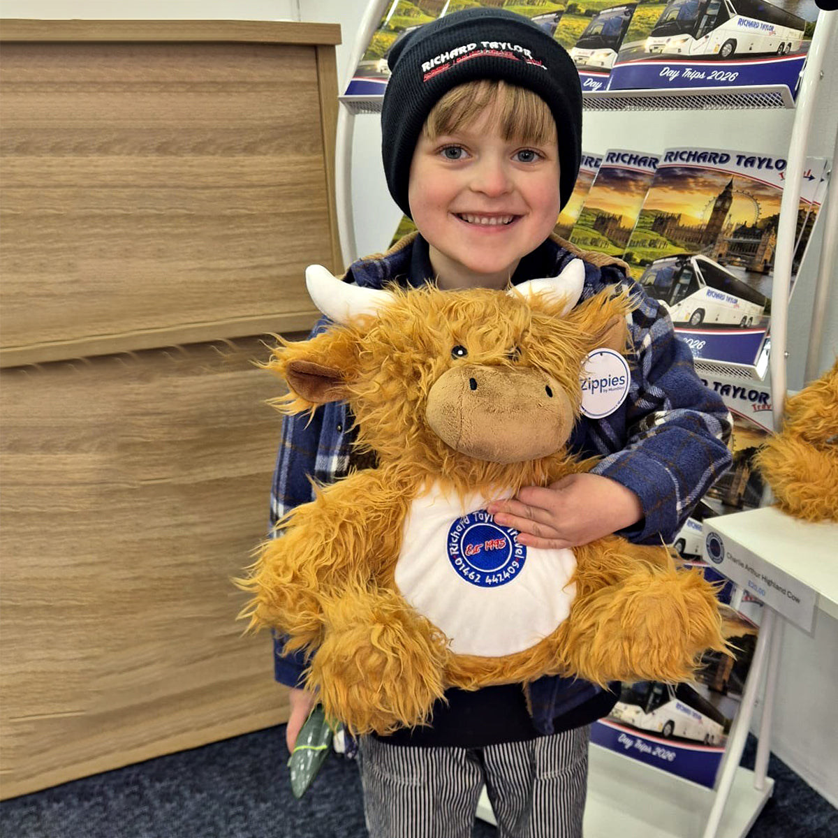 A smiling young boy wearing a blue beanie and blue plaid jacket holds a fluffy golden-brown Charlie Arthur Highland cow plush toy with a white belly patch. The scene is the Richard Taylor Travel shop which books coach day trips and tours