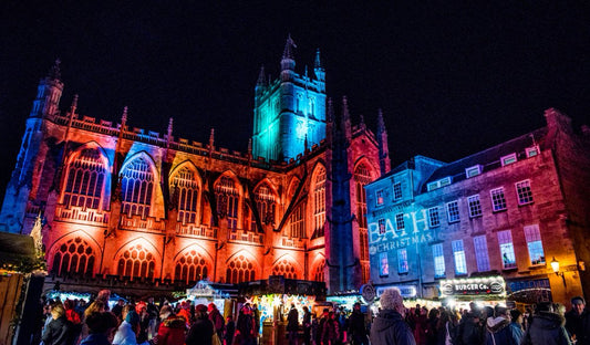 Bath Cathedral illuminated with colourful lights at night, surrounded by people and stalls at the Christmas Market.