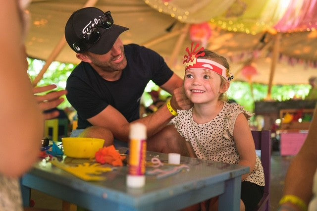Man and young girl sitting at a table making head bands with colourful decorations in an outdoor setting at BeWILDerwood. BeWILDerwood is an award-winning, whimsical outdoor woodland adventure park for families, with locations in England. Based on books by author Tom Blofeld, it offers imaginative, nature-based play, including treetop mazes, wobbly wires, slippery slides, boat rides, and character-driven storytelling for children aged 2 to 12.