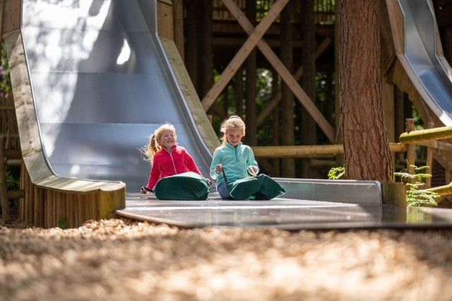 Two children playing on a playground slide at BeWILDerwood. BeWILDerwood is an award-winning, whimsical outdoor woodland adventure park for families, with locations in England. Based on books by author Tom Blofeld, it offers imaginative, nature-based play, including treetop mazes, wobbly wires, slippery slides, boat rides, and character-driven storytelling for children aged 2 to 12.