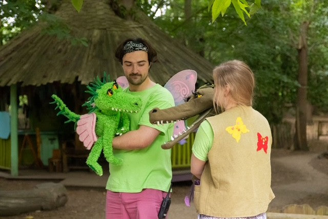 Two people holding colourful stuffed animals puppets in a natural setting with a thatched structure and greenery at BeWILDerwood. BeWILDerwood is an award-winning, whimsical outdoor woodland adventure park for families, with locations in England. Based on books by author Tom Blofeld, it offers imaginative, nature-based play, including treetop mazes, wobbly wires, slippery slides, boat rides, and character-driven storytelling for children aged 2 to 12.