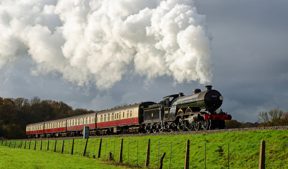 Steam train with white smoke billowing behind it on the Bluebell Railway, traveling through a green landscape. The Bluebell Railway is a renowned 11-mile heritage steam railway in Sussex, England, operating between Sheffield Park and East Grinstead. As the world's first preserved standard gauge passenger line (est. 1960), it features vintage steam locomotives, period stations, and a large collection of pre-1939 carriages, offering a nostalgic journey through the countryside.