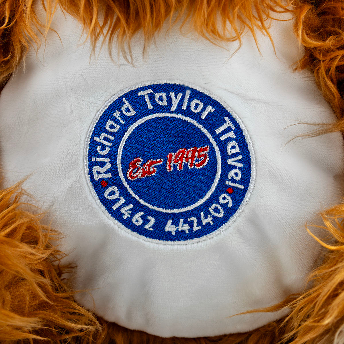 A close-up of a white plush cushion or pet bed with a circular embroidered patch in the center. The patch is blue with white text reading 'Richard Taylor Travel' and 'Est 1995.