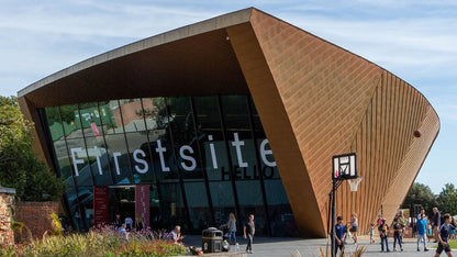 The Firstsite Learning Centre building with its distinctive curved terracotta-colored wooden facade and large glass entrance under a blue sky, with students walking in front.