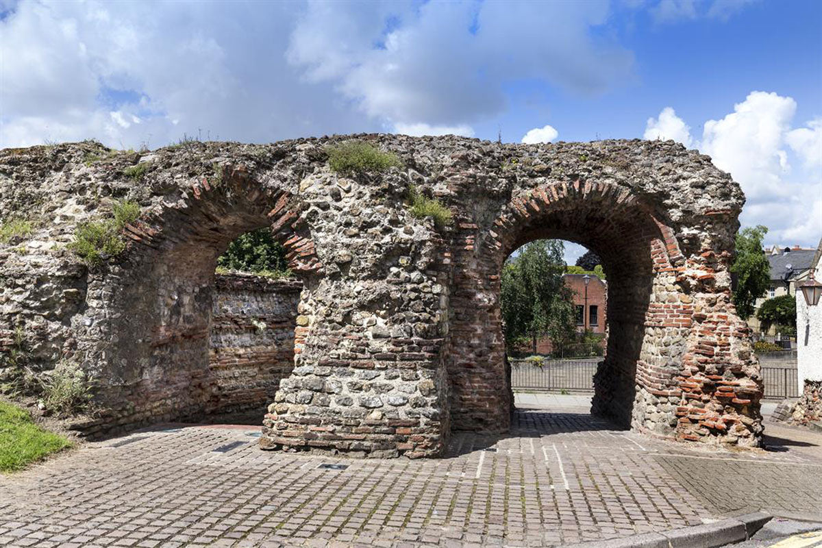 Ancient Roman stone archway ruins with brick detailing under a blue sky with white clouds, featuring a paved stone walkway in the foreground in Colchester.