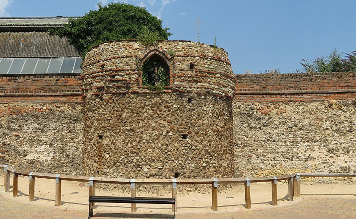 An outdoor stone cylindrical roman ruin in Colchester with a small arched window, set against a brick wall under a blue sky. A wooden bench and low railing are in the foreground.