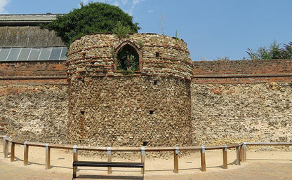 An outdoor stone cylindrical roman ruin in Colchester with a small arched window, set against a brick wall under a blue sky. A wooden bench and low railing are in the foreground.