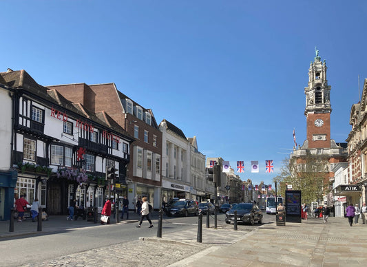 A sunny street scene in a historic town of Colchester with timber-framed buildings, pedestrians, cars, and a tall clock tower under a clear blue sky.
