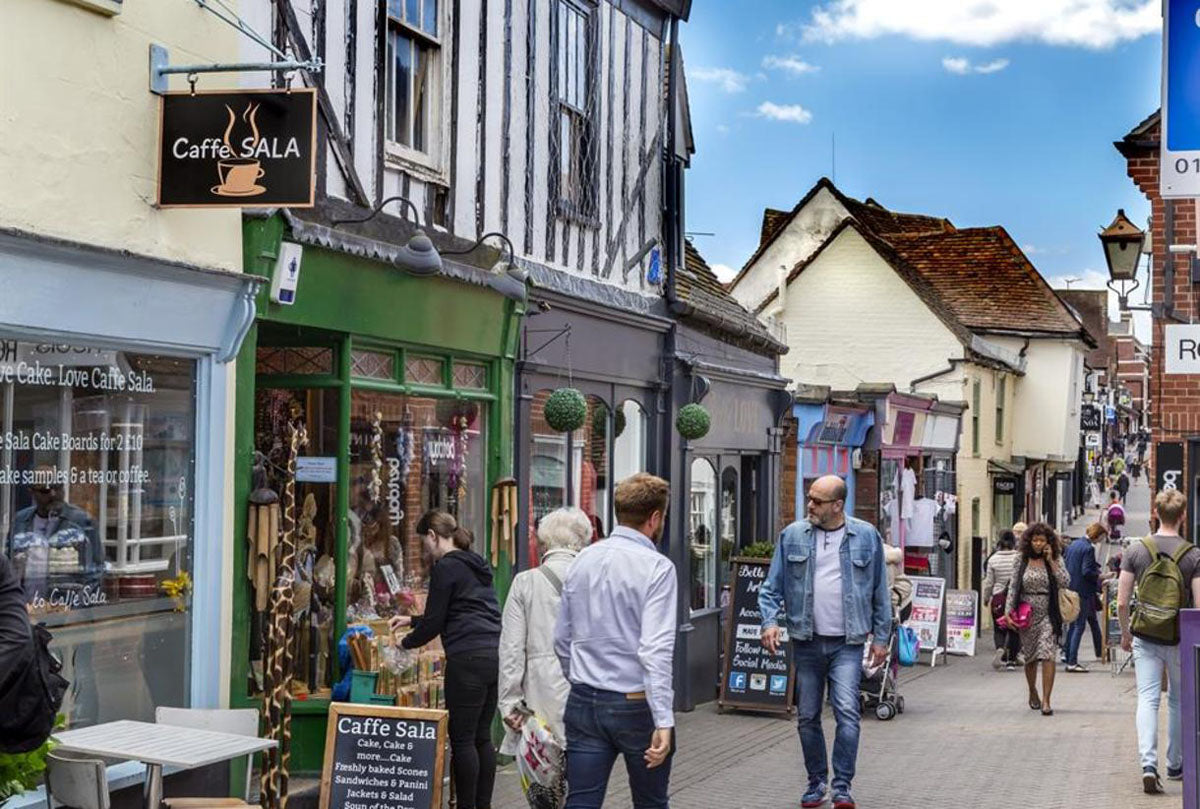 A bustling outdoor street scene in front  in Colchester of Caffe Sala, showing pedestrians walking past a shop with a green facade and timber-framed buildings under a blue sky.