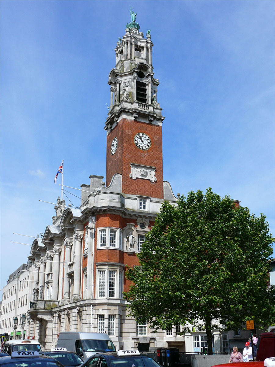 A tall brick and stone clock tower building under a blue sky in Colchester, with a large green tree in the foreground and a few cars and pedestrians at the bottom.