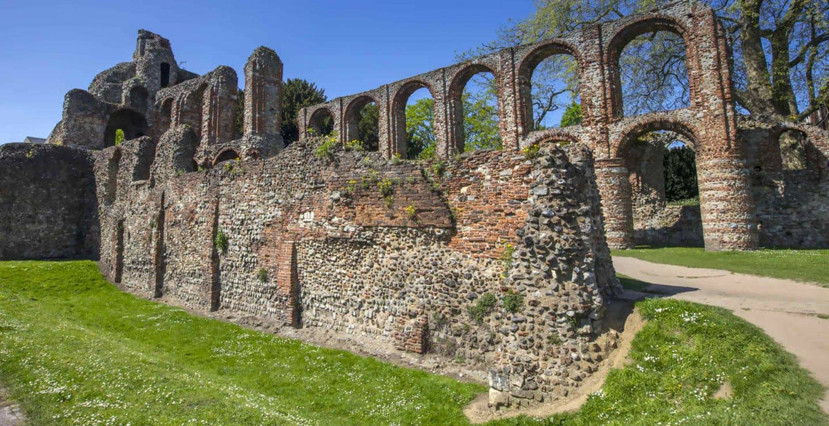 Colchester Stone ruins with arches and walls under a blue sky, with green grass and a dirt path in the foreground.