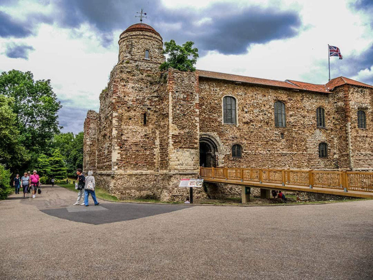 Colchester outdoor scene showing a large historic stone building with a round tower, arched entrance, wooden walkway, paved foreground, trees, and a cloudy sky. A few people are walking near the building, and a flag flies on the roof.