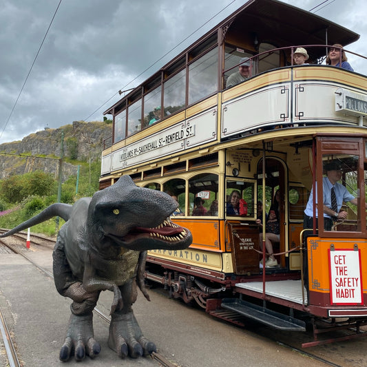 Vintage trolley car at dinosaur Special Day on tracks against a mountainous background at Crich Tramway. Crich Tramway Village, located in Derbyshire, is an open-air museum and home to the National Tramway Museum, featuring a restored period village street and a working mile-long tramway. Visitors can ride vintage electric trams through scenic woodland. The site includes a workshop, exhibitions, a pub, and a woodland walk.