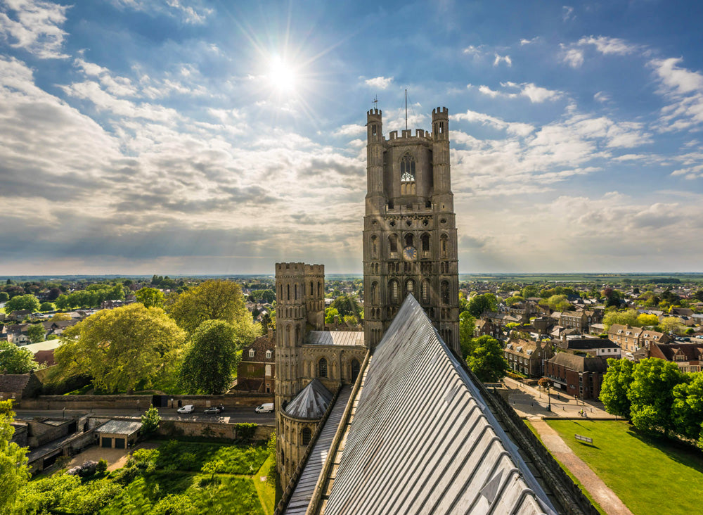 Ely Cathedral aerial view of the surrounding town and greenery under a blue sky. Ely Cathedral, known as the "Ship of the Fens," is a stunning 11th-century Romanesque and Gothic masterpiece in Cambridgeshire, famed for its unique Octagon Tower, long nave, and vibrant stained glass. The compact city centre, surrounding the cathedral, features historic streets, Oliver Cromwell’s House, and a bustling market, offering a charming mix of medieval history and modern amenities.