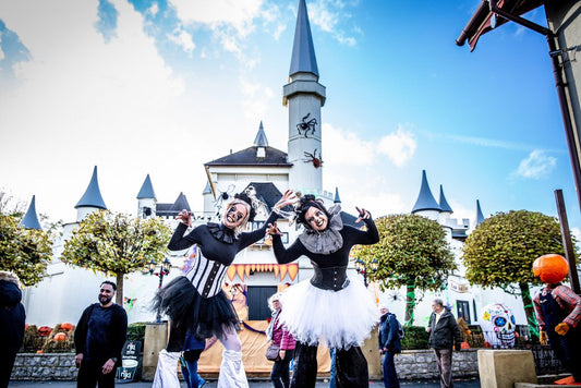 Two performers in black and white Halloween costumes dancing in front of a castle-like structure with trees and people in the background.