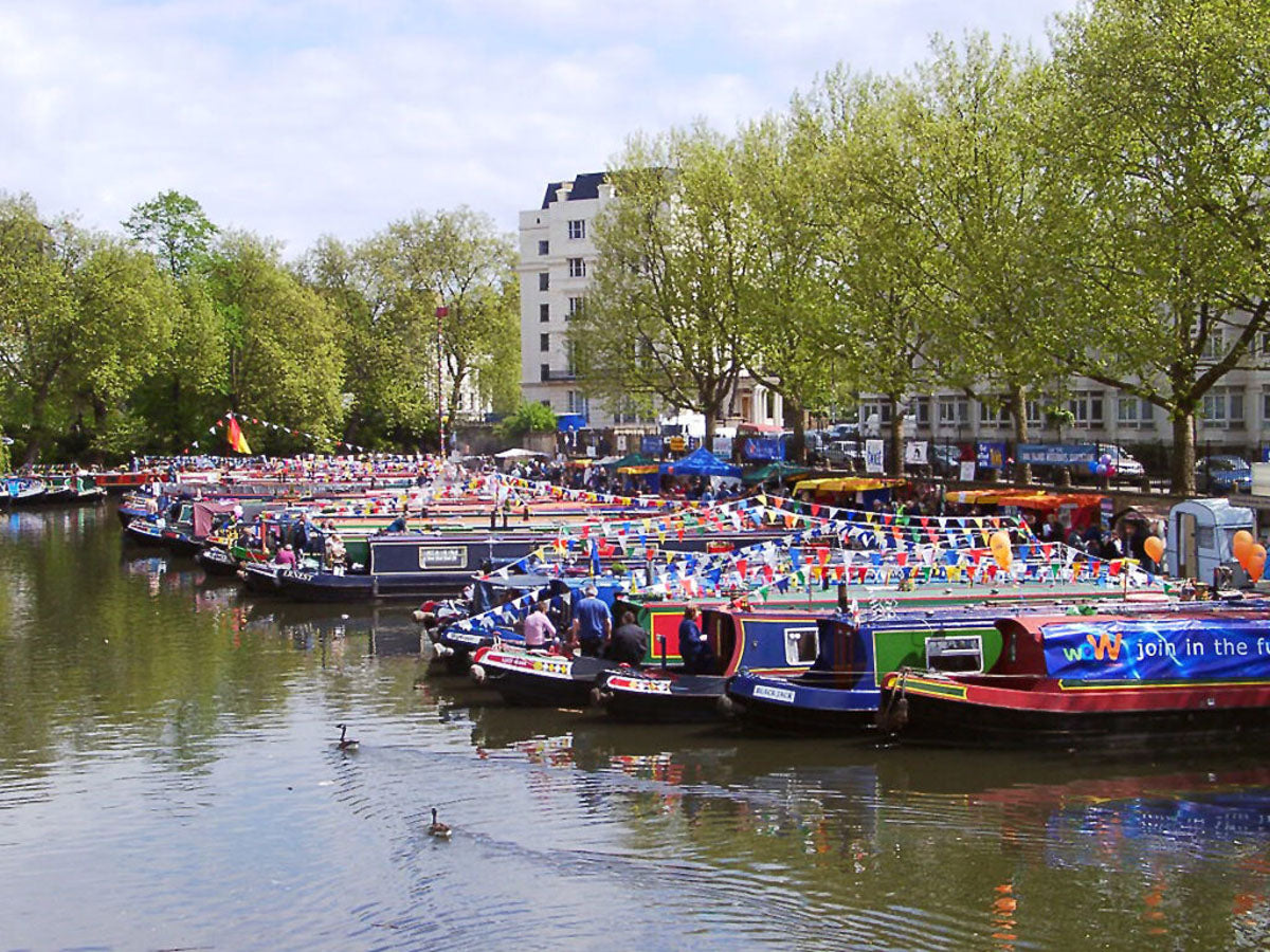 IWA Canalway Festival canal scene with many colorful narrowboats docked along the water, surrounded by green trees and buildings under a partly cloudy sky.