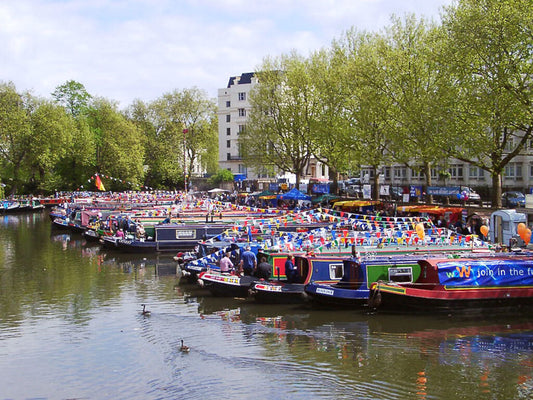 IWA Canalway Festival canal scene with many colorful narrowboats docked along the water, surrounded by green trees and buildings under a partly cloudy sky.