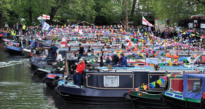 IWA Canalway Festival canal scene with many narrowboats docked along the water, decorated with colorful flags and bunting, with people standing on and around the boats. Trees line the background.