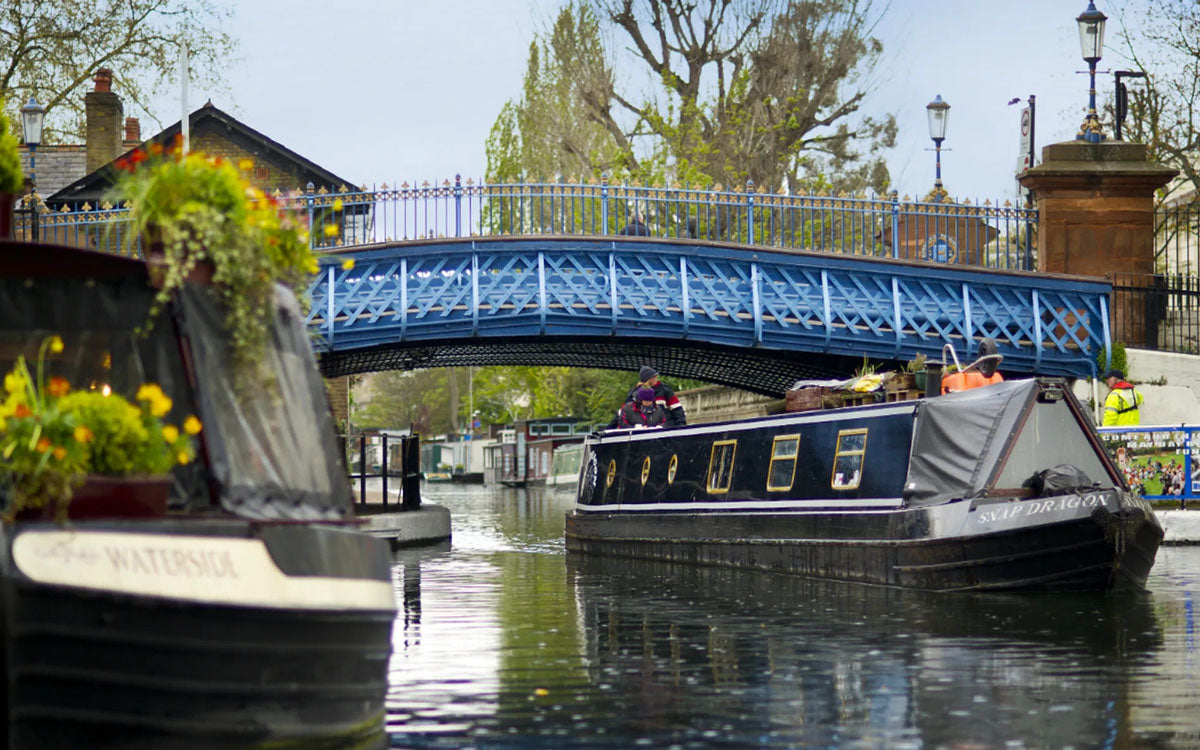 IWA Canalway Festival narrowboat on a canal passing under a blue bridge, with other boats and trees in the background.