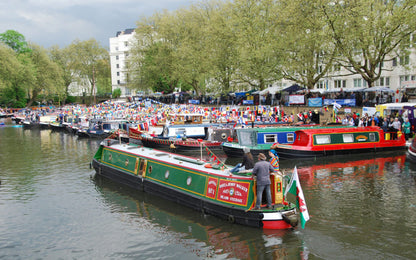 IWA Canalway Festival  green and red narrowboat with a person standing on the back deck is photographed on a canal, with other boats, trees, and a crowd along the riverbank in the background.