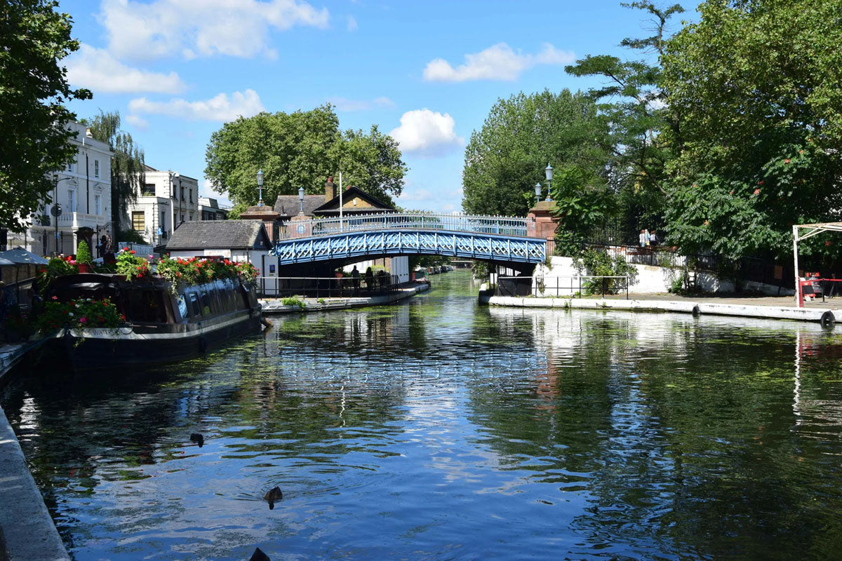 IWA Canalway Festival scenic view of a canal with a blue bridge, surrounded by lush green trees, boats, and buildings under a bright blue sky with white clouds.