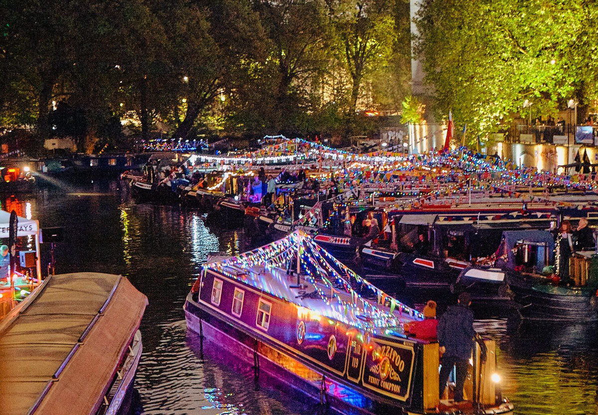 IWA Canalway Festival nighttime canal scene with several narrowboats decorated with bright string lights. Trees and buildings line the canal, and many people are visible on and around the boats.