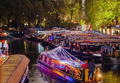 IWA Canalway Festival nighttime canal scene with several narrowboats decorated with bright string lights. Trees and buildings line the canal, and many people are visible on and around the boats.