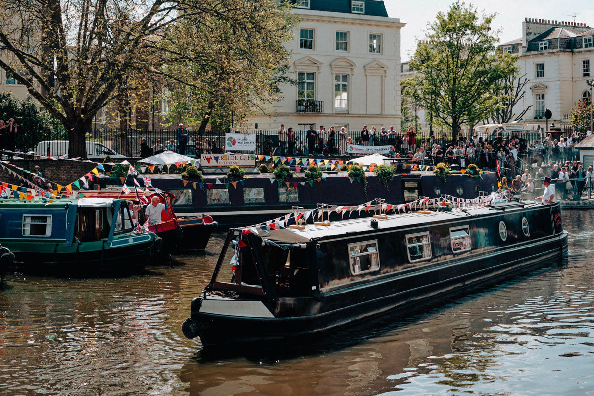 IWA Canalway Festival black narrowboat with colorful bunting floats on a canal, surrounded by other boats, onlookers, trees, and a light-colored building in the background.