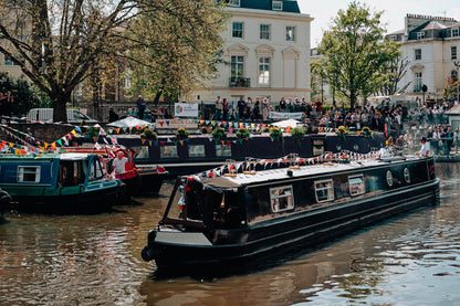 IWA Canalway Festival black narrowboat with colorful bunting floats on a canal, surrounded by other boats, onlookers, trees, and a light-colored building in the background.