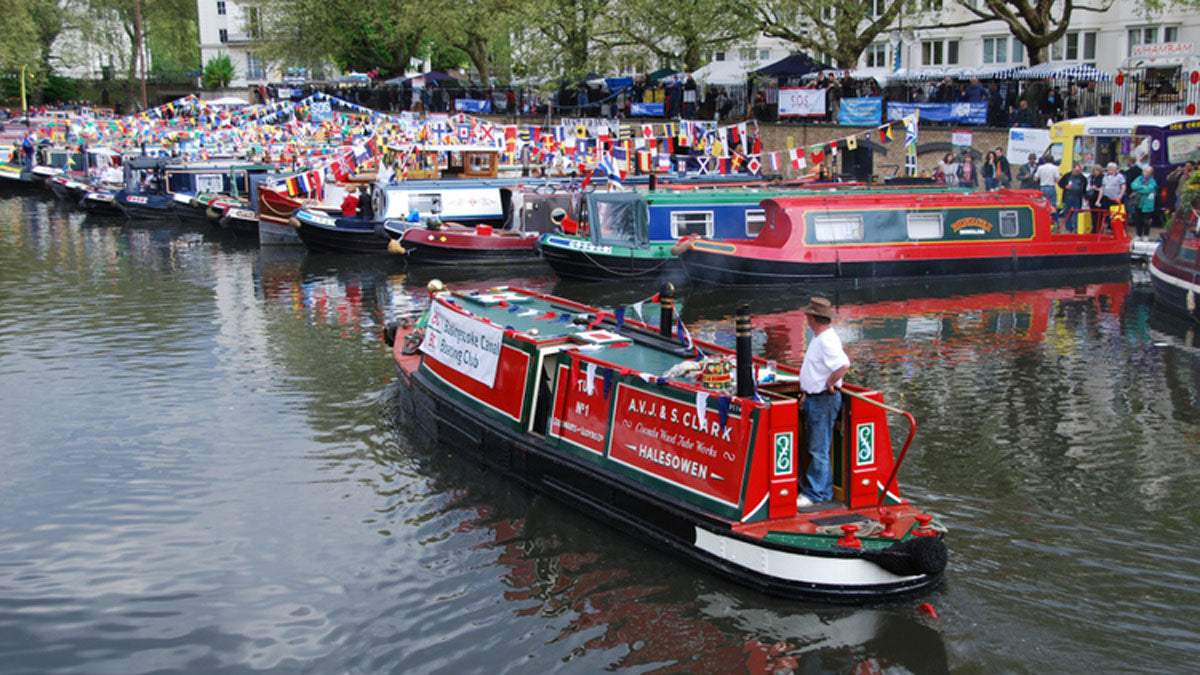 IWA Canalway Festival  red and black narrowboat navigating a canal lined with many other boats, flags, and onlookers.