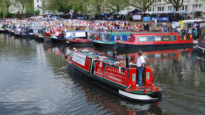 IWA Canalway Festival  red and black narrowboat navigating a canal lined with many other boats, flags, and onlookers.