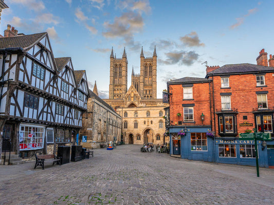 Lincoln historic town square with a large cathedral in the background, framed by traditional timber-framed houses on the left and red brick buildings on the right under a blue sky with light clouds.