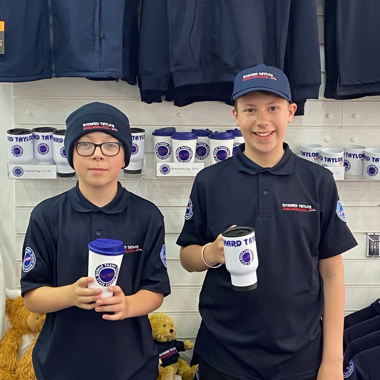 Two young men wearing matching Richard Taylor Travel dark navy polo shirts and baseball caps with 'Richard Taylor Travel' branding, holding white and blue branded tumblers in what appears to be a retail store setting.