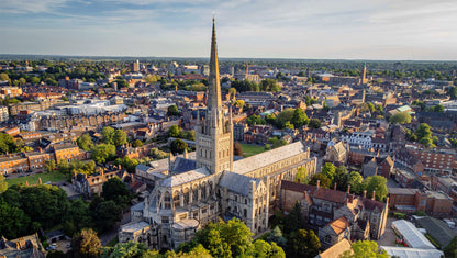 Aerial view of Norwich Cathedral with a tall spire surrounded by a cityscape. Norwich Cathedral, founded in 1096, is a premier example of Romanesque architecture in Europe, featuring a 315-foot spire (second tallest in England), the largest monastic cloisters in the country, and over 1,000 medieval roof bosses. Located in Norfolk, this 900-year-old landmark combines Norman, Gothic, and Perpendicular styles with a notable vaulted ceiling.