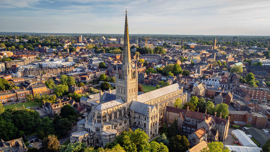 Aerial view of Norwich Cathedral with a tall spire surrounded by a cityscape. Norwich Cathedral, founded in 1096, is a premier example of Romanesque architecture in Europe, featuring a 315-foot spire (second tallest in England), the largest monastic cloisters in the country, and over 1,000 medieval roof bosses. Located in Norfolk, this 900-year-old landmark combines Norman, Gothic, and Perpendicular styles with a notable vaulted ceiling.