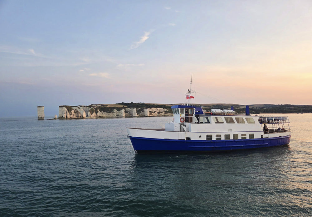 Boat on water with a sunset and Old Harry Rocks are a famous series of white chalk sea stacks and stumps located at Handfast Point, on the Isle of Purbeck in Dorset, southern England, near Poole and Studland Bay. They mark the most eastern point of the Jurassic Coast, in the background