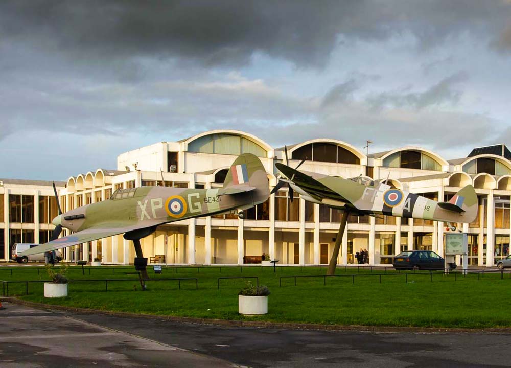 Vintage Spitfire airplanes on display in front of RAF Hendon with a cloudy sky. The Royal Air Force Museum London (RAF Hendon) is a free, family-friendly museum in North London, located on the historic former London Aerodrome. It features over 100 aircraft across multiple hangars, interactive galleries, and exhibits documenting the history of aviation, the Battle of Britain, and the RAF’s centenary.