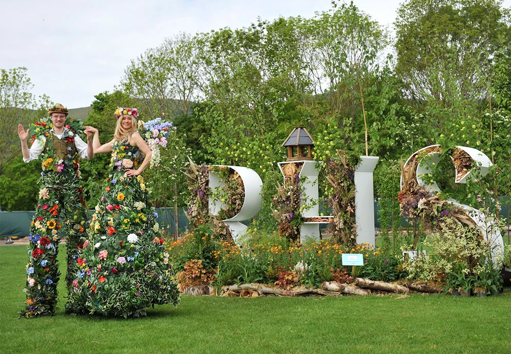 Two people in floral outfits standing in a garden with decorative floral letters spelling 'RHS' at RHS Malvern. The RHS Malvern Spring Festival is a premier annual horticultural event held at the Three Counties Showground, Worcestershire, marking the start of the summer gardening season. It features renowned show gardens, a vast Plant Village, floral marquees, and celebrity experts like Monty Don. The festival offers food, shopping, and family activities in a scenic setting.