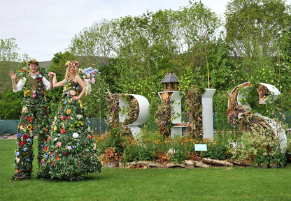Two people in floral outfits standing in a garden with decorative floral letters spelling 'RHS' at RHS Malvern. The RHS Malvern Spring Festival is a premier annual horticultural event held at the Three Counties Showground, Worcestershire, marking the start of the summer gardening season. It features renowned show gardens, a vast Plant Village, floral marquees, and celebrity experts like Monty Don. The festival offers food, shopping, and family activities in a scenic setting.
