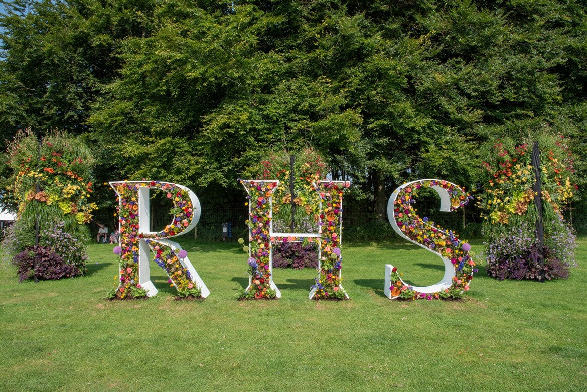 Floral letters spelling 'RHS' on a grassy area in the Sandringham Estate with trees in the background. Sandringham Estate is a 20,000-acre Norfolk country estate and private home to British monarchs since 1862. Famed as the Royal Family's Christmas retreat, it features Sandringham House (circa 1870), 60 acres of gardens, the St Mary Magdalene Church, and extensive public woodland. The estate is a working, sustainable landscape within the Norfolk Coast Area of Outstanding Natural Beauty. 