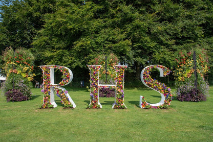 Floral letters spelling 'RHS' on a grassy area in the Sandringham Estate with trees in the background. Sandringham Estate is a 20,000-acre Norfolk country estate and private home to British monarchs since 1862. Famed as the Royal Family's Christmas retreat, it features Sandringham House (circa 1870), 60 acres of gardens, the St Mary Magdalene Church, and extensive public woodland. The estate is a working, sustainable landscape within the Norfolk Coast Area of Outstanding Natural Beauty. 