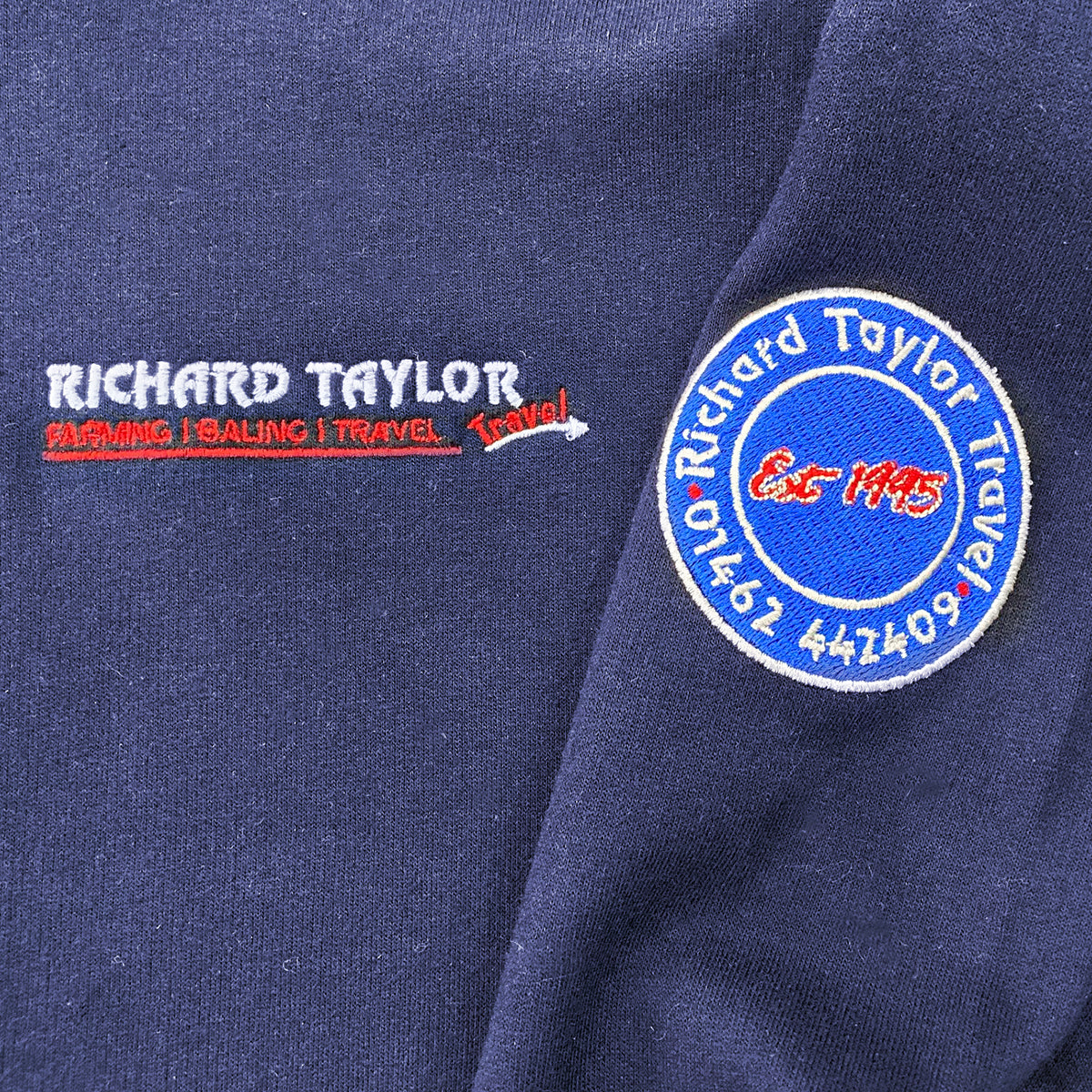Close-up of a navy blue fabric surface showing embroidered text and a circular patch. The embroidery reads "RICHARD TAYLOR TRAVEL" with smaller text below in white and red, and the round patch also says "Richard Taylor Travel" with "Est 1995".