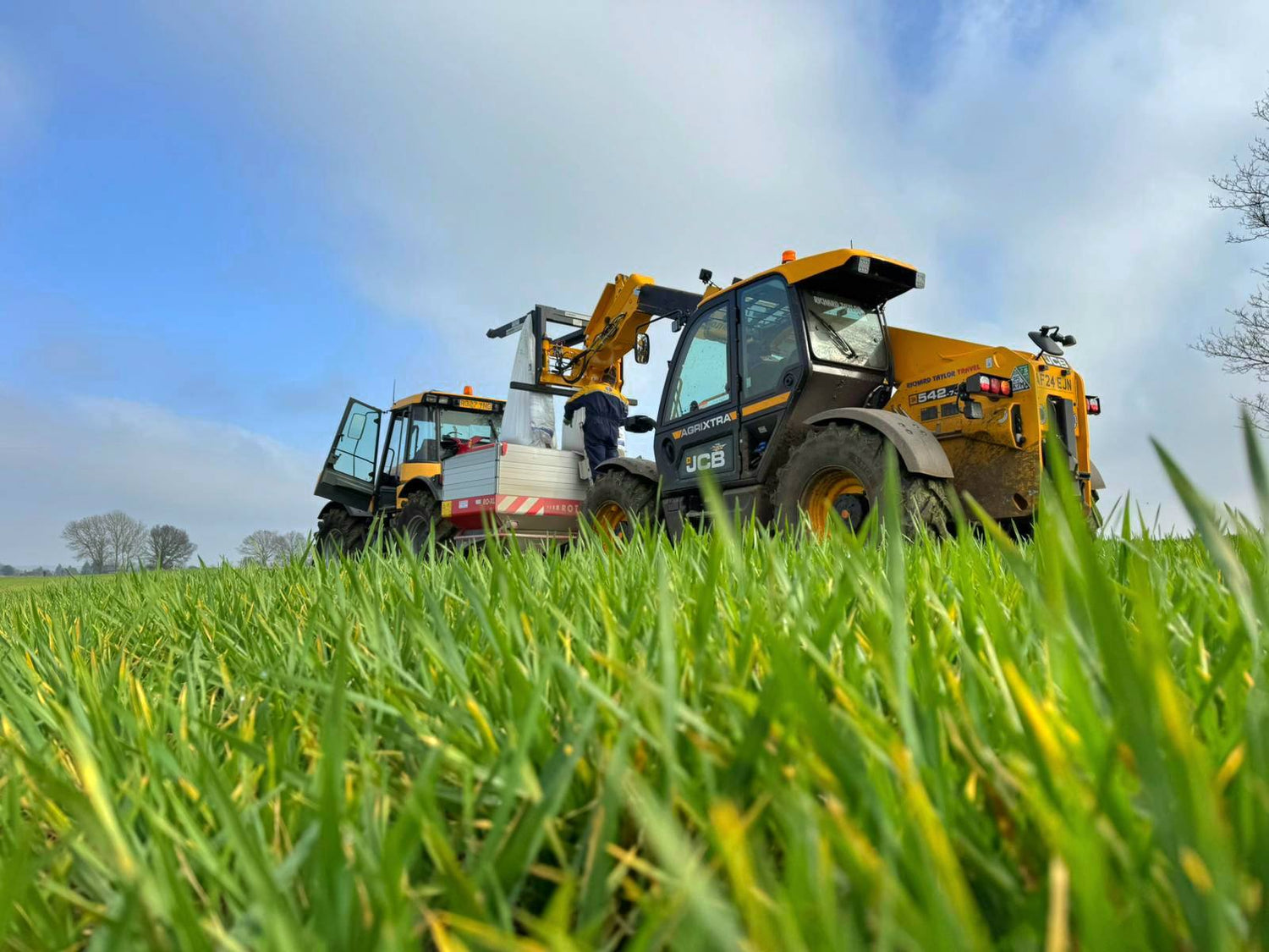 Richard Taylor Travel JCB handler filling tractor spreader vehicles on a grassy field with a blue sky.