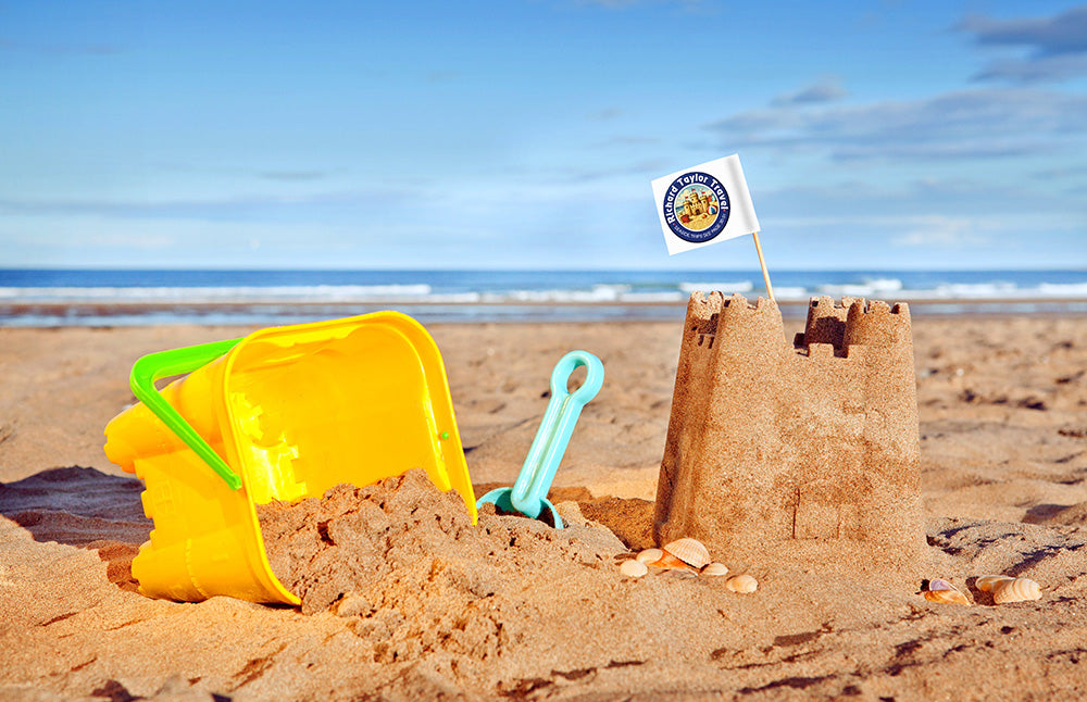 Sandcastle with a Richard Taylor Travel flag, yellow bucket, and blue shovel on a sandy beach with ocean and sky in the background.