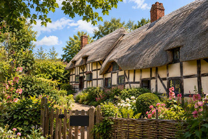 A picturesque thatched cottage with cream walls and dark timber framing, surrounded by a lush garden with pink flowers, a wooden fence, and a wicker basket, under a blue sky with white clouds.