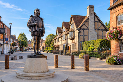 A bronze statue of a historical figure on a stone pedestal stands in the center of a paved town square, with timber-framed houses, brick buildings, bollards, and a blue sky in the background.