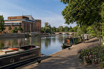 A scenic view of a canal with boats docked along the shore, a brick building on the left, and lush green trees on the right under a blue sky.