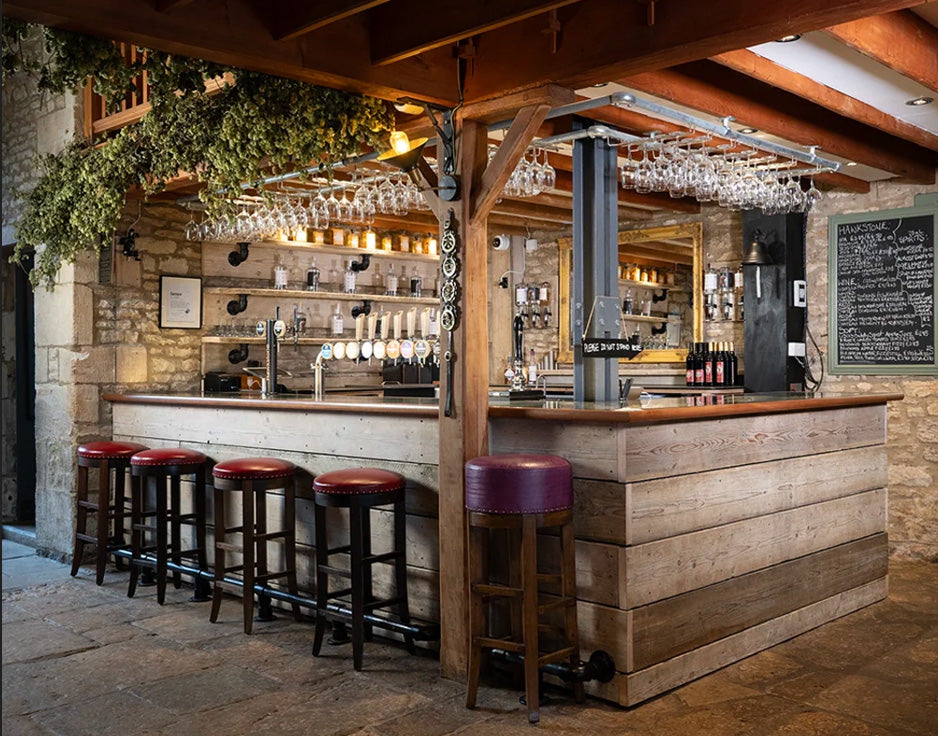 Wooden bar with stools in the Farmers Dog pub. The Farmer's Dog, located in Asthall near Burford in the Cotswolds, is a traditional British pub opened by Jeremy Clarkson in 2024. It focuses exclusively on 100% British-sourced food and drink—including Hawkstone beer—featuring a, butchers, and farm shop. The dog-friendly venue features rustic décor and a lively, rural atmosphere.