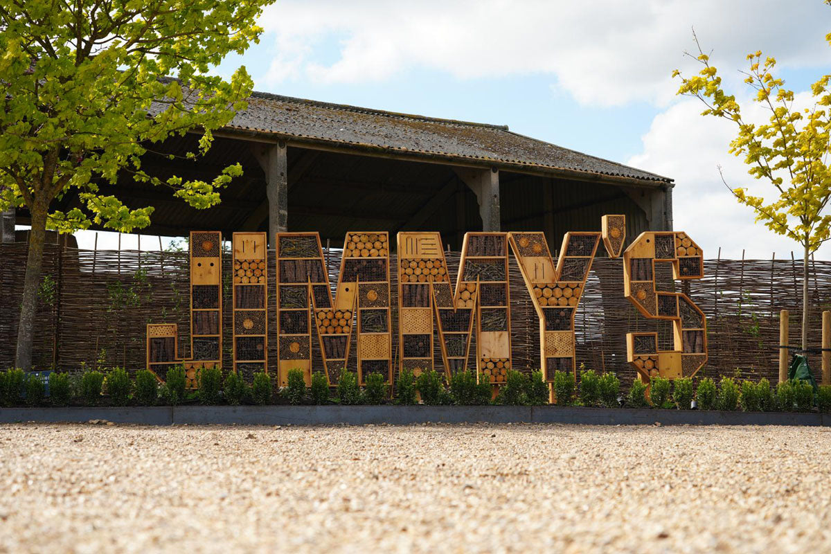Large wooden lettering spelling 'JIMMY's' mounted outdoors in front of a rustic wooden pavilion, with gravel ground in the foreground and trees on both sides at Jimmy's Farm and Wildlife Park entrance.